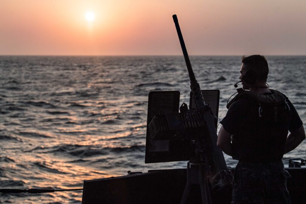 A U.S. Navy sailor stands watch at a gun station aboard USS Jason Dunham at sunset while transiting the Bab al-Mandeb Strait