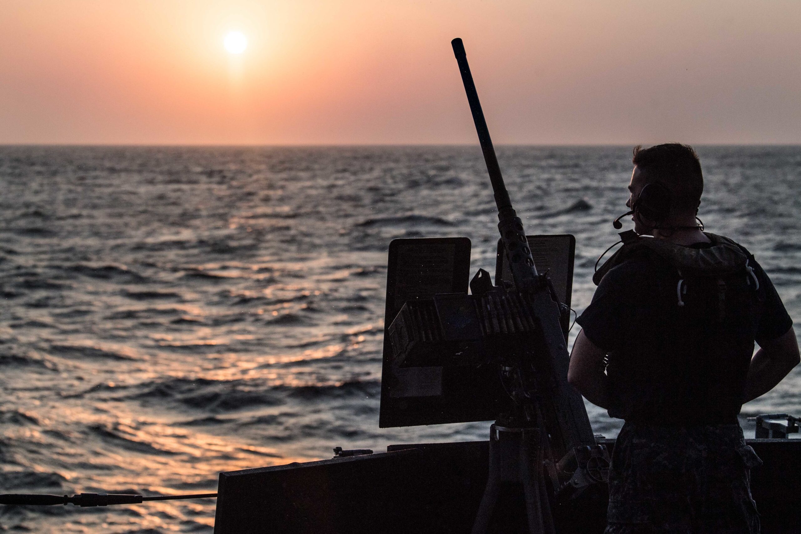 A U.S. Navy sailor stands watch at a gun station aboard USS Jason Dunham at sunset while transiting the Bab al-Mandeb Strait